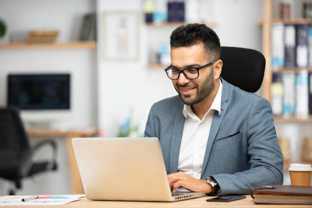 A man in a suit focused on his laptop, immersed in work at a modern office setting.