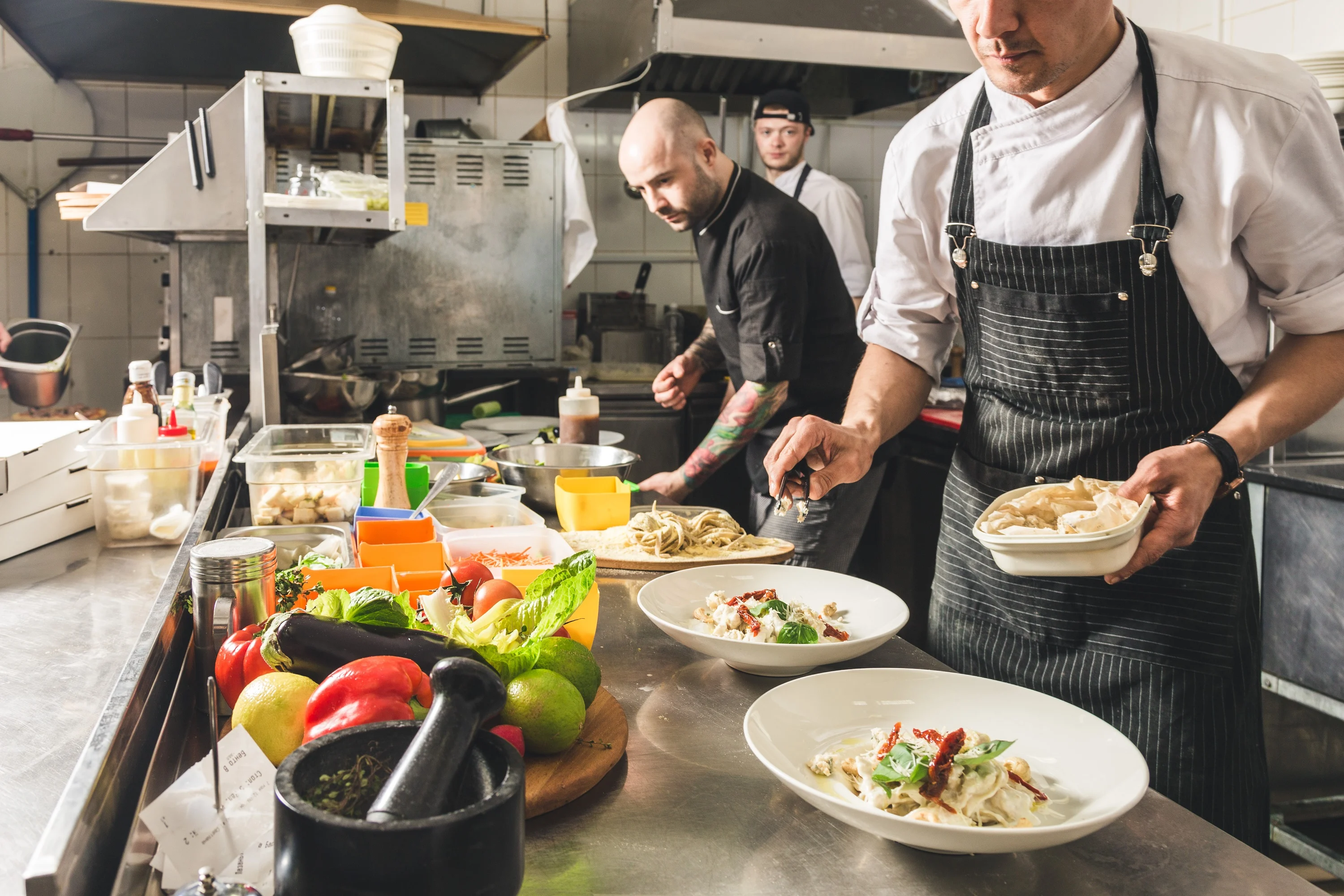 Chefs in a kitchen preparing plates of pasta, with fresh ingredients like peppers and limes on the counter.