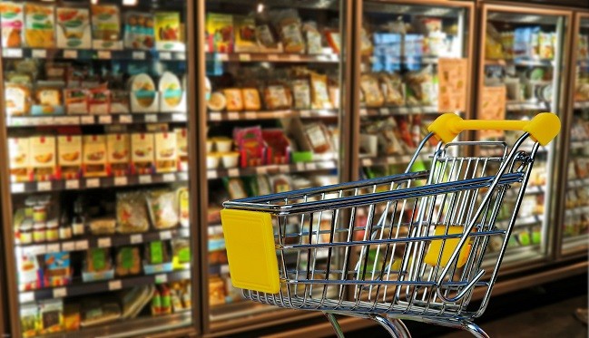 Empty shopping cart in front of a refrigerated section filled with packaged goods in a grocery store.