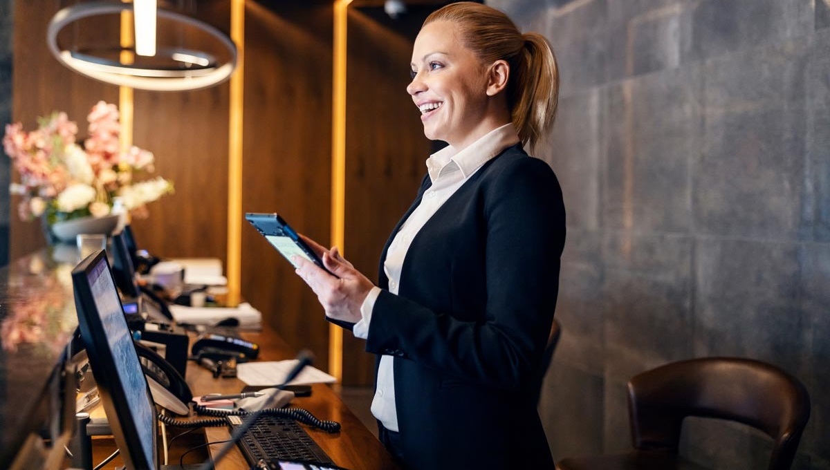 A professional woman in a dark suit stands at a stylish reception desk, holding a tablet, with blurred office equipment in the background.