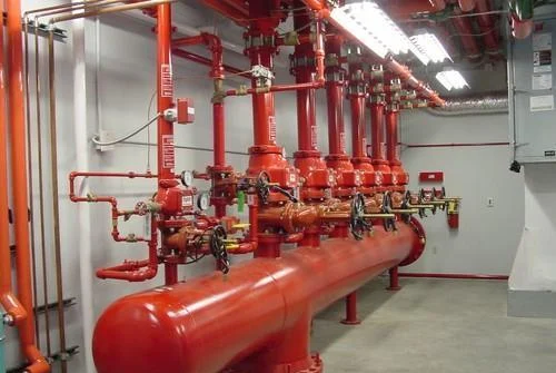 A row of red industrial pipes and valves against a gray wall, with overhead fluorescent lighting illuminating the workspace.