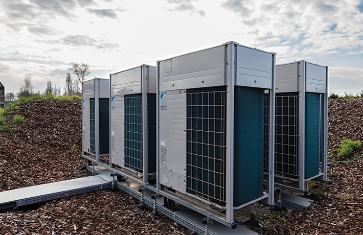 Four large air conditioning units are positioned on a metal platform over a bed of mulch, with a cloudy sky in the background.